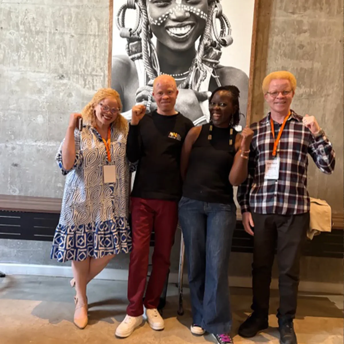 Four people standing together indoors and smiling in front of a large black-and-white portrait photograph.