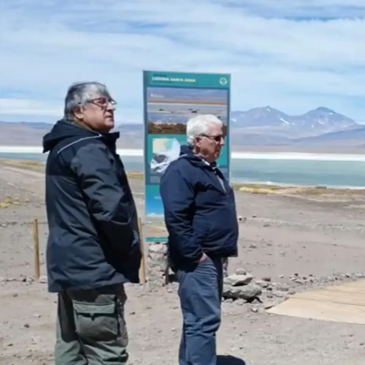 Two people standing outdoors beside an informational sign overlooking a mountainous landscape and lake.