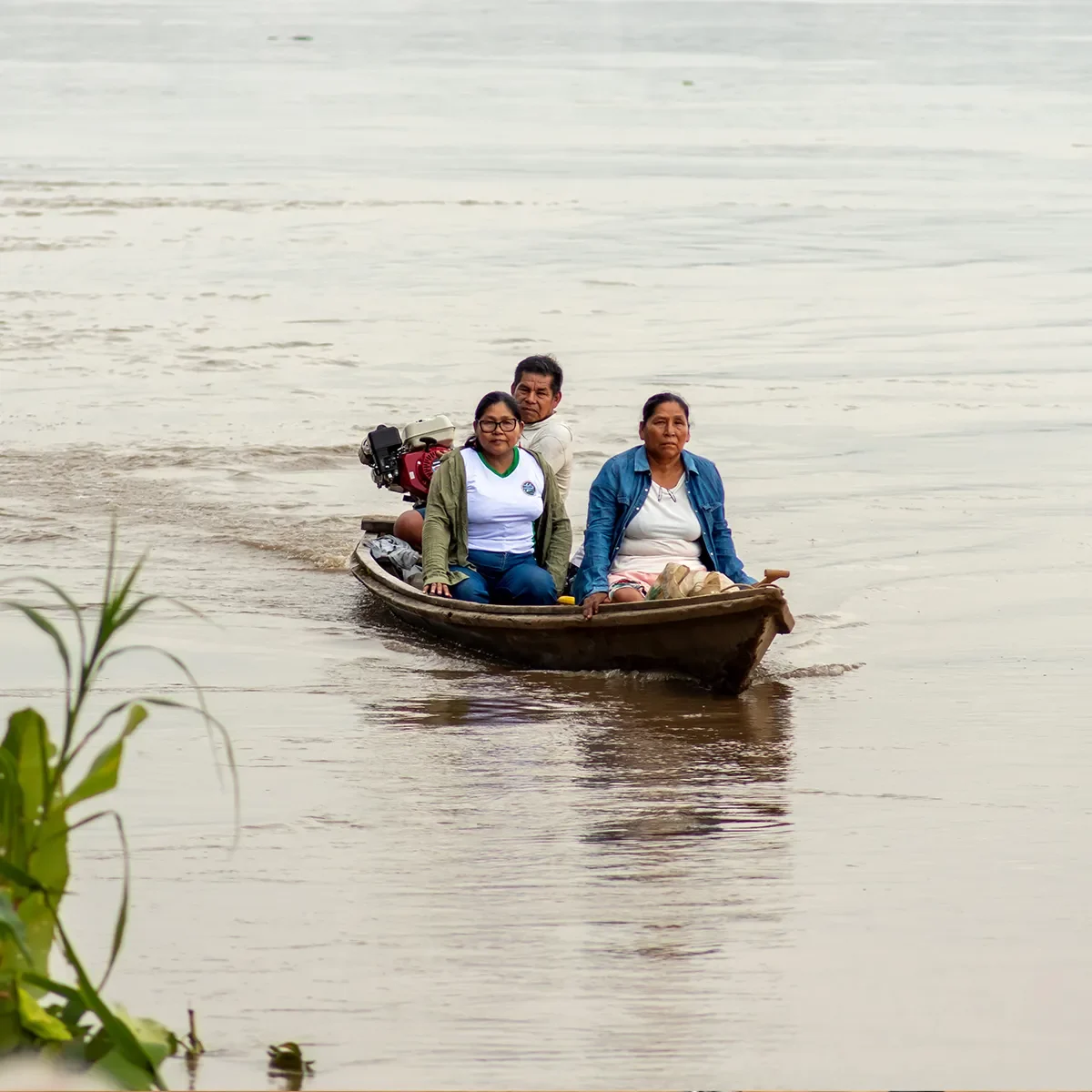 Three people travelling by small wooden boat across a wide river or lake in a rural landscape.