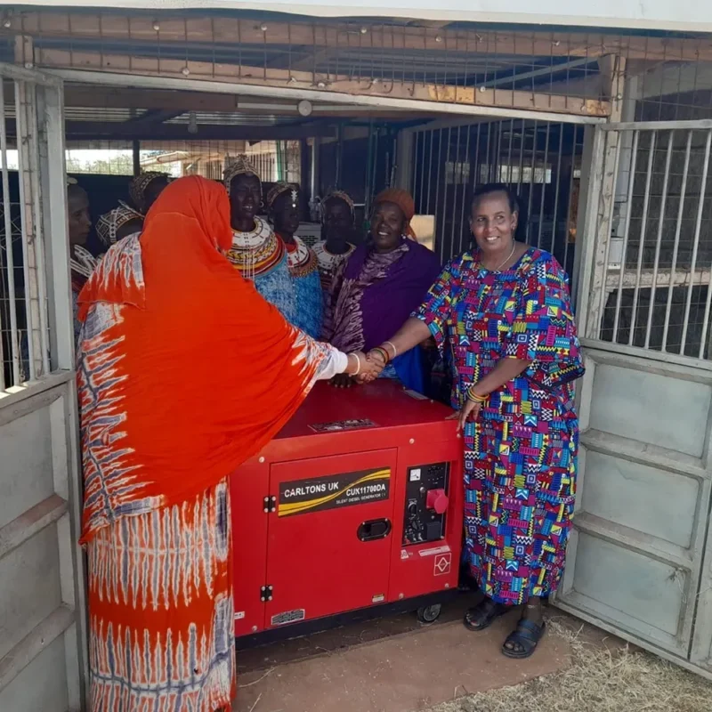 Group of women gathered around a red generator inside a community structure, with two women shaking hands during a community handover or partnership moment.