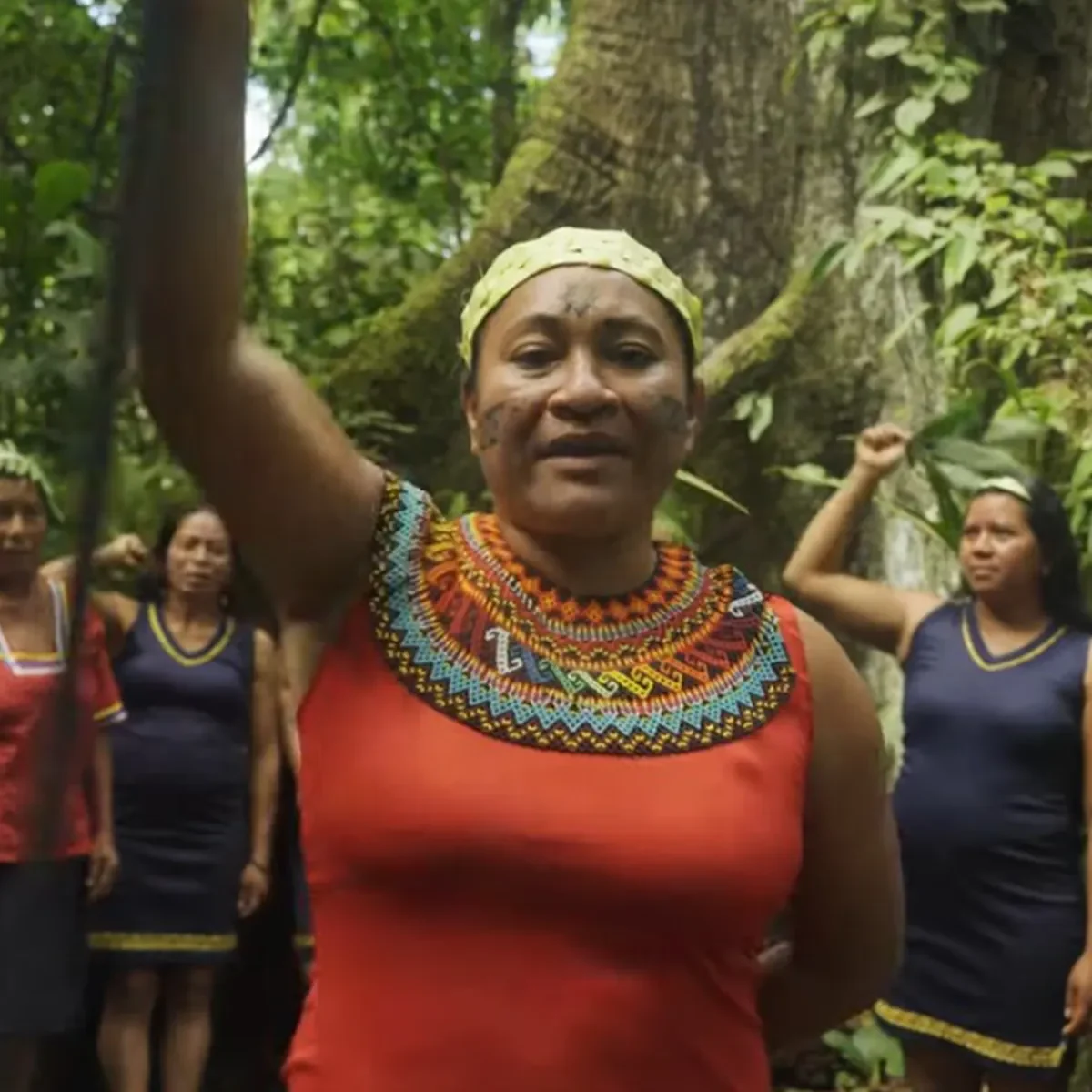 Indigenous woman wearing colourful traditional beadwork raises her arm while speaking outdoors, with community members gathered in the background.